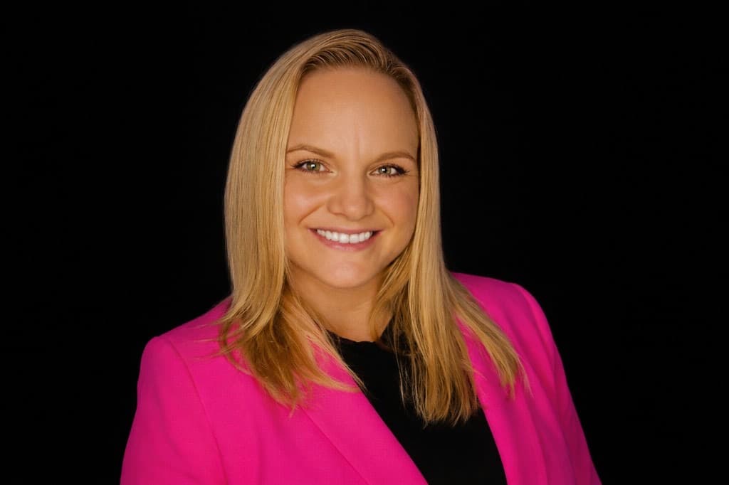 Professional headshot of Michelle, founder of Biz Boost Agency, in a bright pink blazer against a black background.